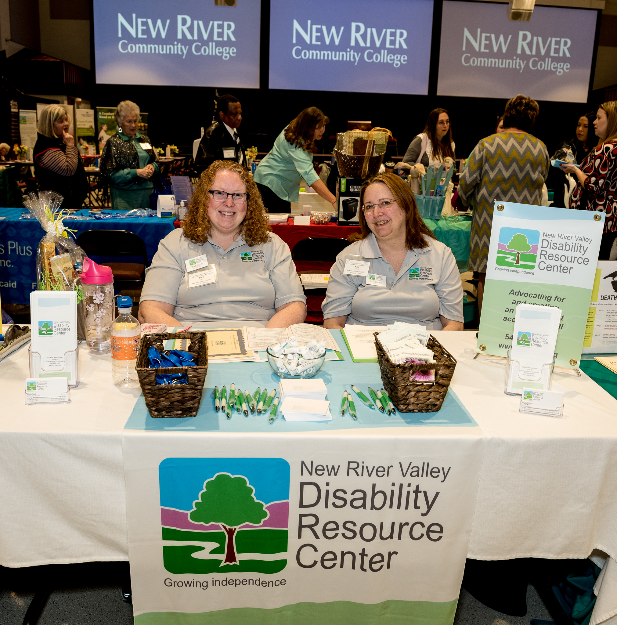 Two smiling white women are seated at an exhibition table in the middle of a large room. Behind them are large display screens on the wall and also the back of other exhibition tables with visitors to them.