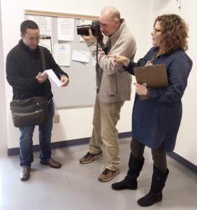 A woman with reddish brown hair speaks to a reporter, a dark haired man looking at a small notepad and writing notes, and a photographer, a tall gray haired man holding a camera to his eye