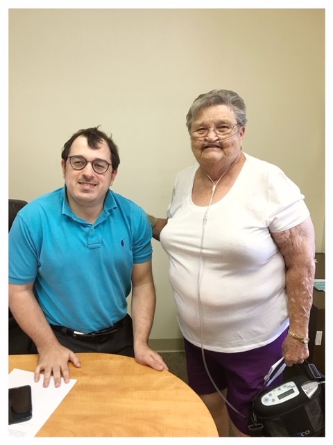 On the left stands Matt Shelor, a 36 year old white make standing roughly 5 feet tall with dark hair and dark framed glasses. On the right stands Ms. Farley, who is an older white female with gray short hair and glasses standing next to Matt. She is wearing a white short-sleeve blouse and purple shorts, is wearing an oxygen cannula and is holding a portable oxygen machine in her left hand. Both of them are smiling at the camera as they stand behind Matt’s desk in his office.