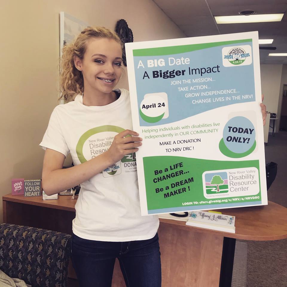 A teenage girl with blond curly hair stands smiling and holding a large sign that highlights the Give Big fundraising event for that day